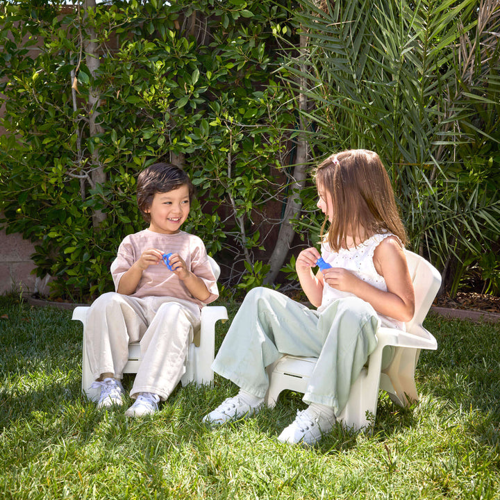 Two kids sitting on the white chairs, on the grass with tree behind them