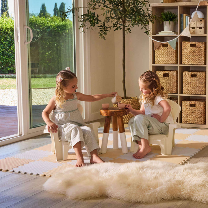 Two girls having a tea party sitting on the chairs, with a small table between them, in a playroom setting with beige walls
