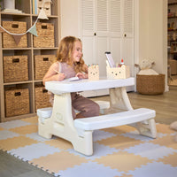 Girl with long blonde hair sitting at the table with pen and paper. Setting is a playroom with shelves and table is sitting on play mats.