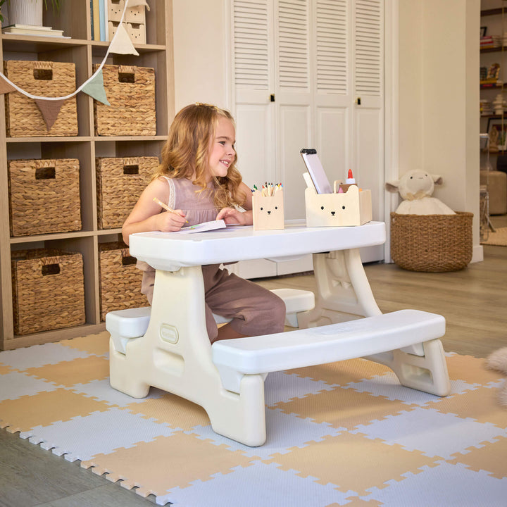 Girl with long blonde hair sitting at the table with pen and paper. Setting is a playroom with shelves and table is sitting on play mats.