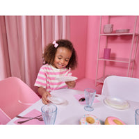 A young girl with curly hair wearing a pink and white striped shirt at a white table in a pink room, holding a white plate. The scene includes pink chairs, shelves, glasses, and toy donuts on the table.