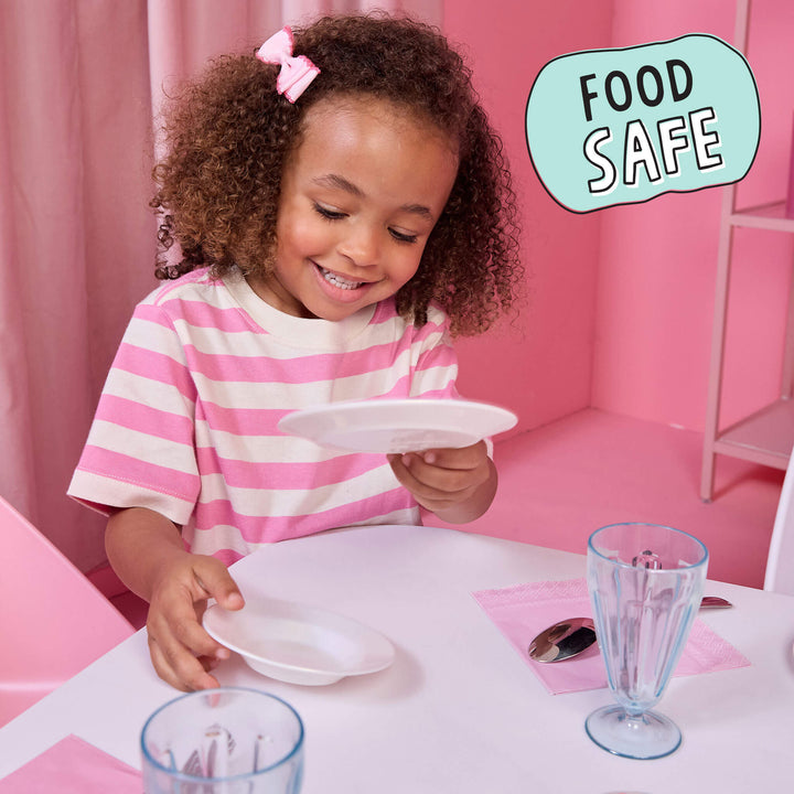 A young girl with curly hair and a pink bow sits at a table in a pink room, holding a small white plate. She is wearing a pink and white striped shirt.